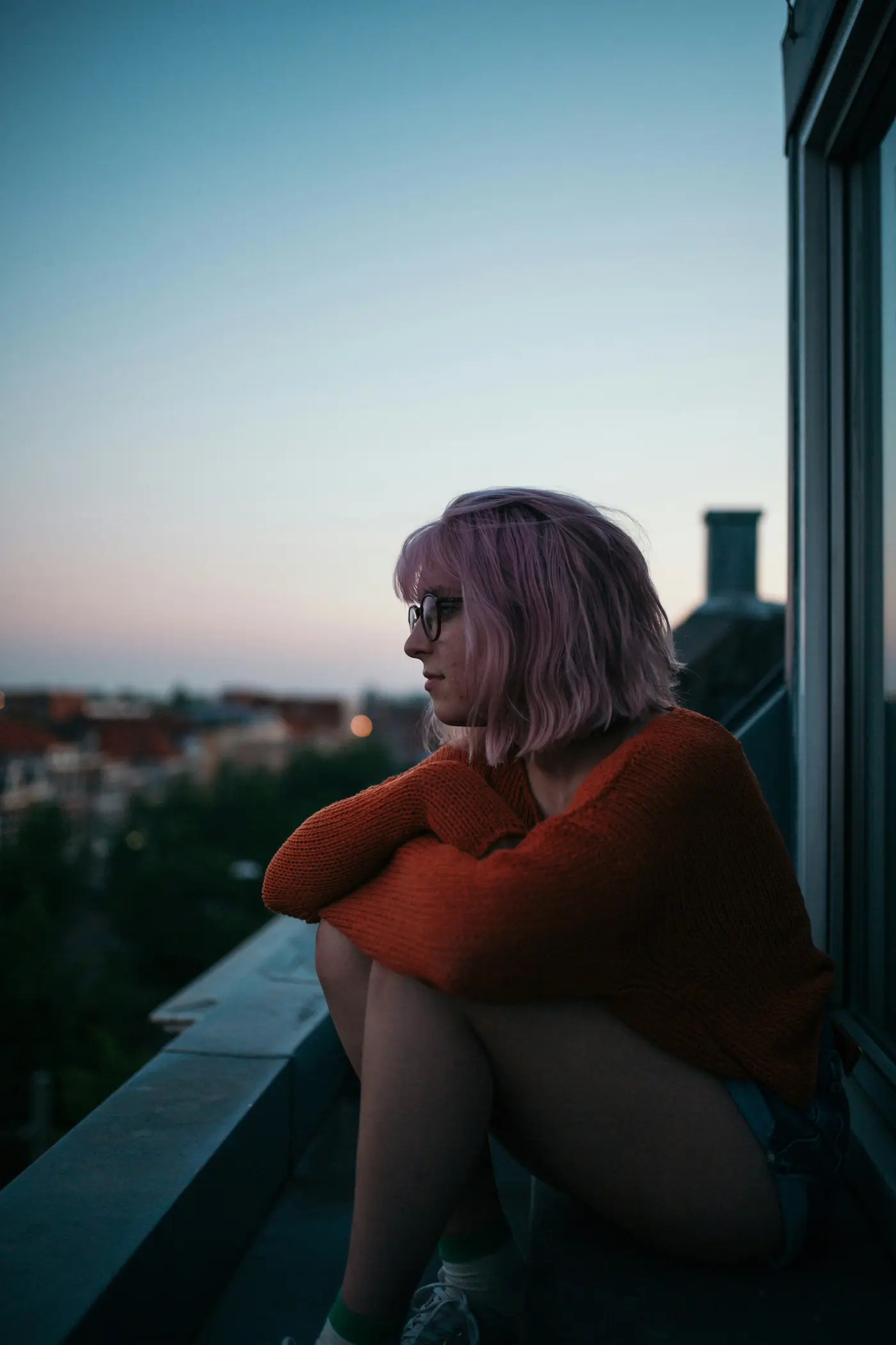 A woman sitting on a balcony at dusk, looking out over the city