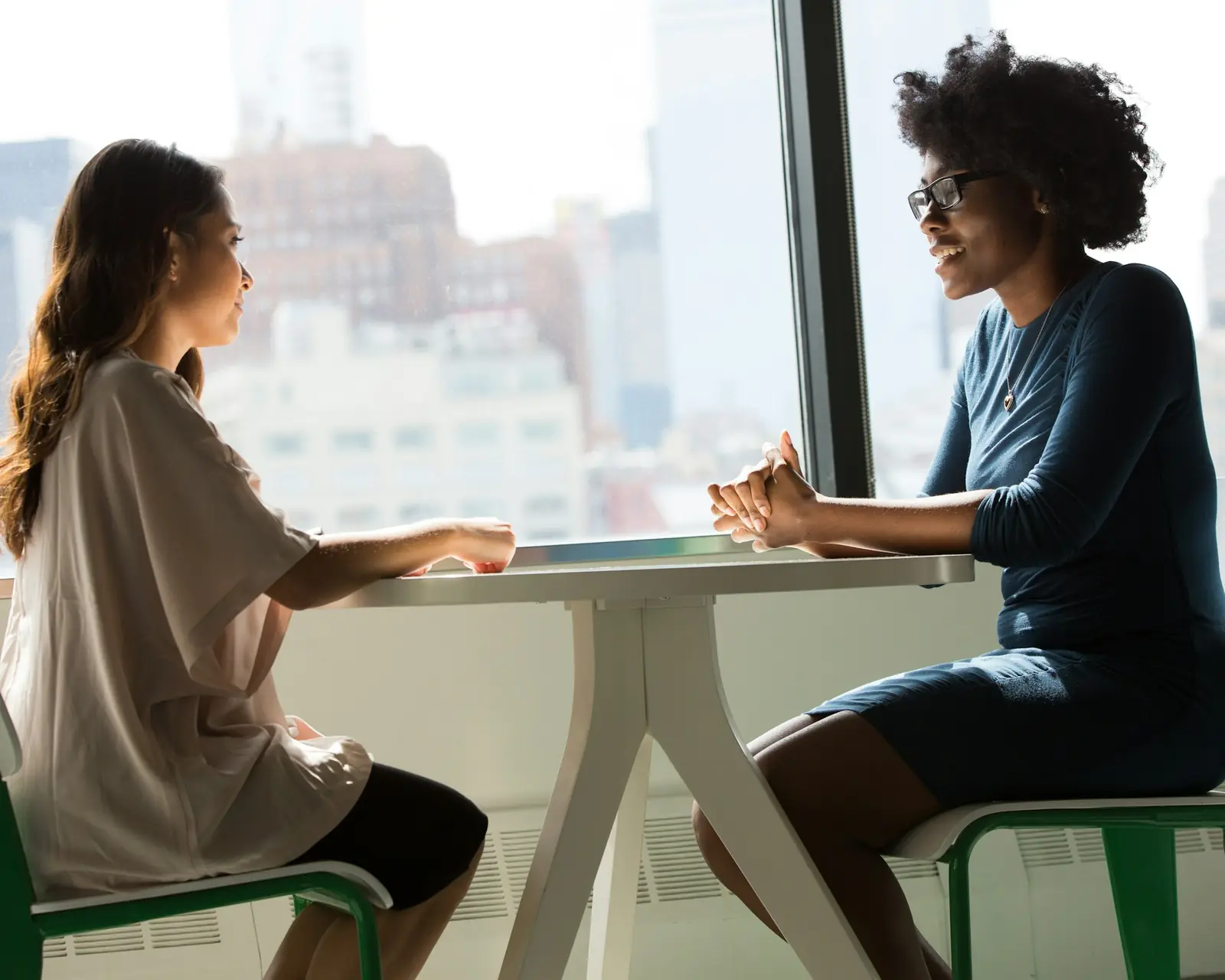 Two women talking at a white table by a window high above the city