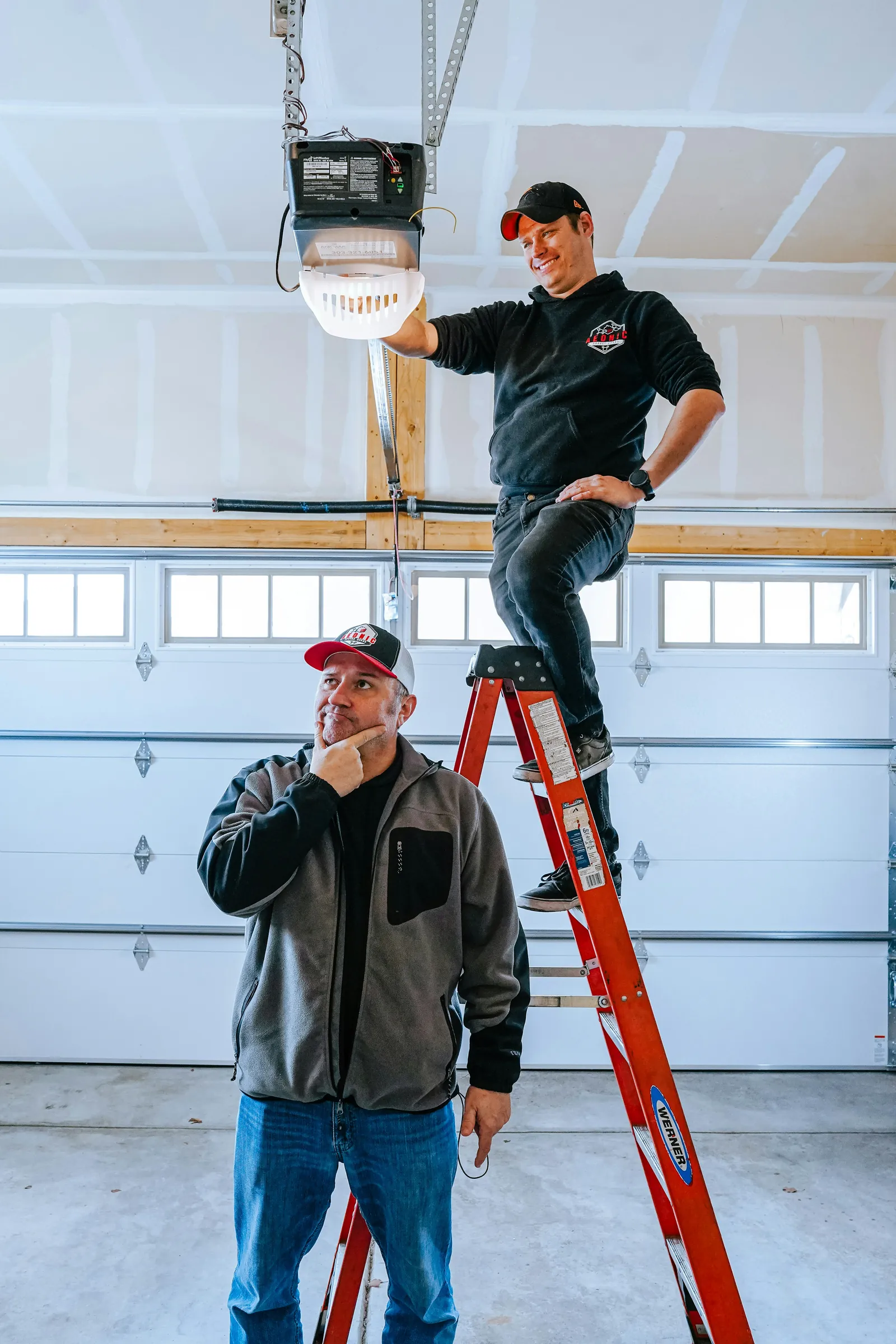 A technician on a ladder installing a garage door opener while the homeowner watches