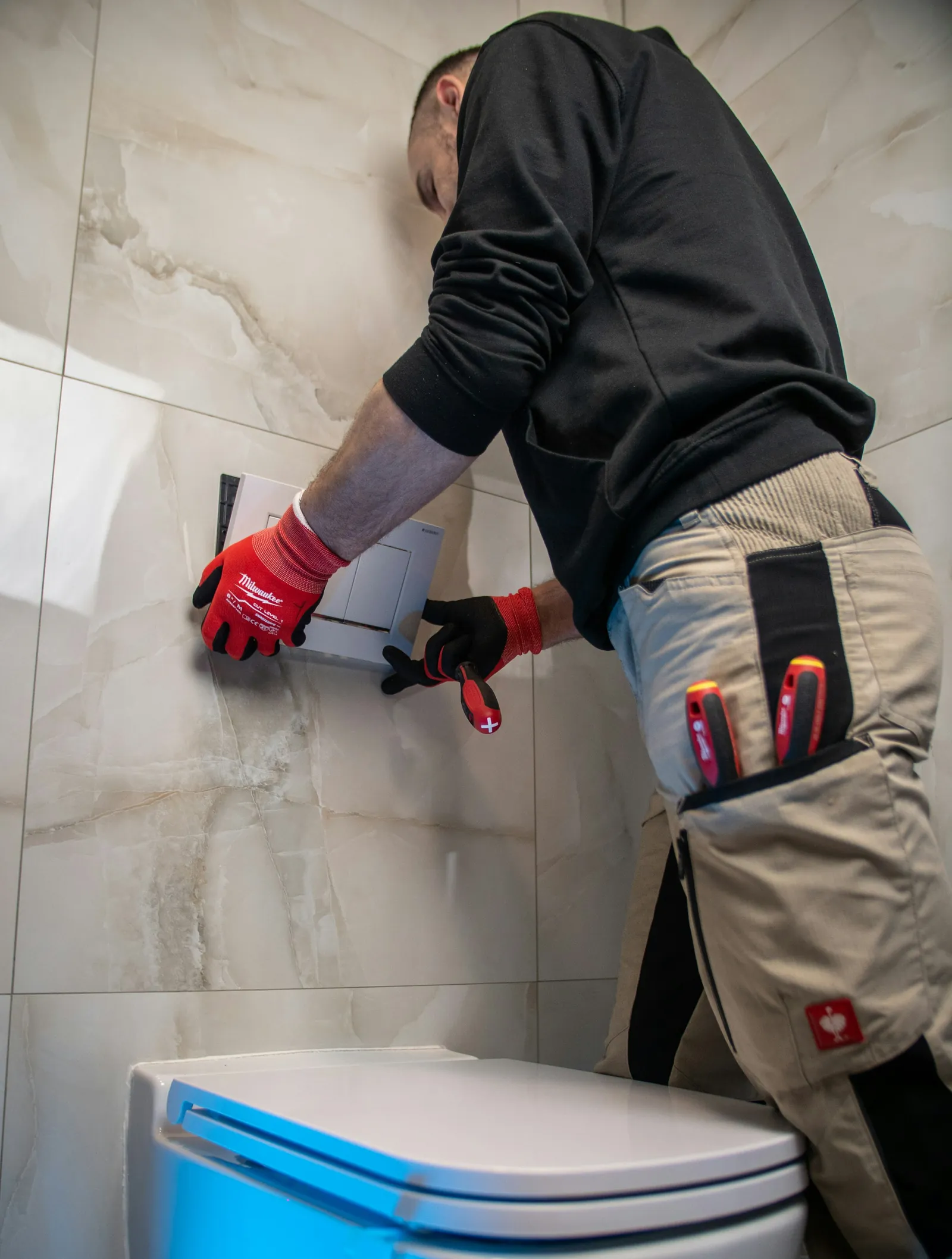 A plumber in work trousers and gloves fitting a pipe behind a bathroom wall