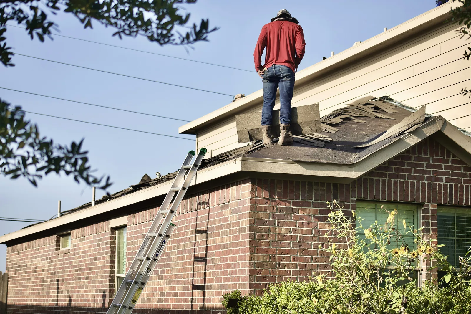 A roofer standing on the roof of a brick house, a ladder against the wall below