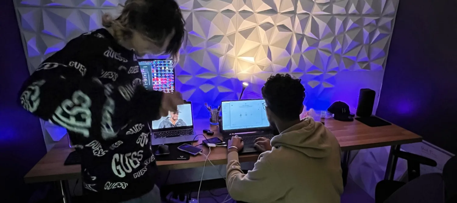 Two of the team at the studio desk under purple light: one leans in over a laptop on a video call while the other types on a second laptop