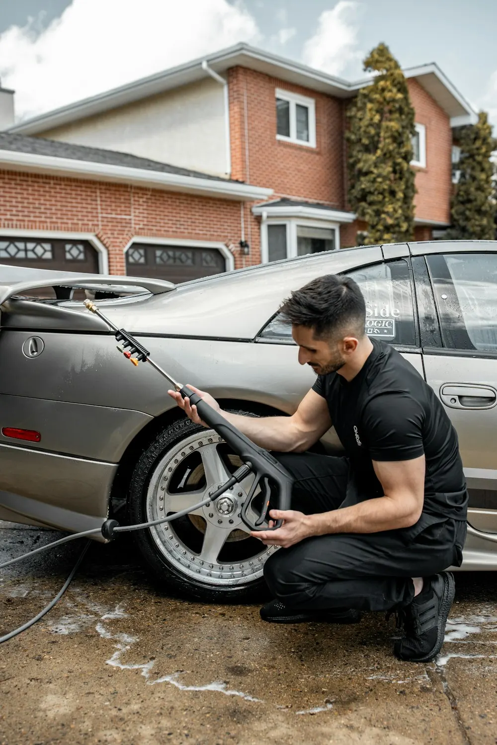 A detailer kneeling at a car's wheel with a pressure washer wand