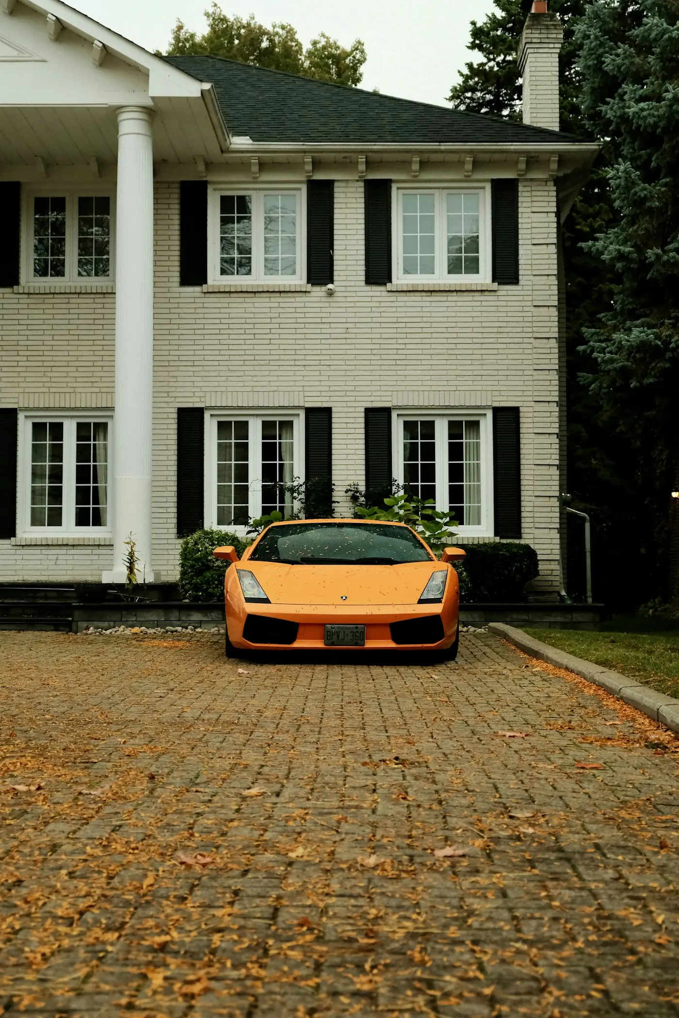 An orange sports car on a clean brick driveway outside a white house